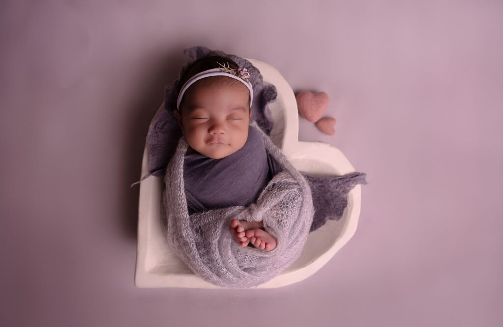 Portrait of newborn wrapped in purple fabric nestled in a heart shaped clay bowl