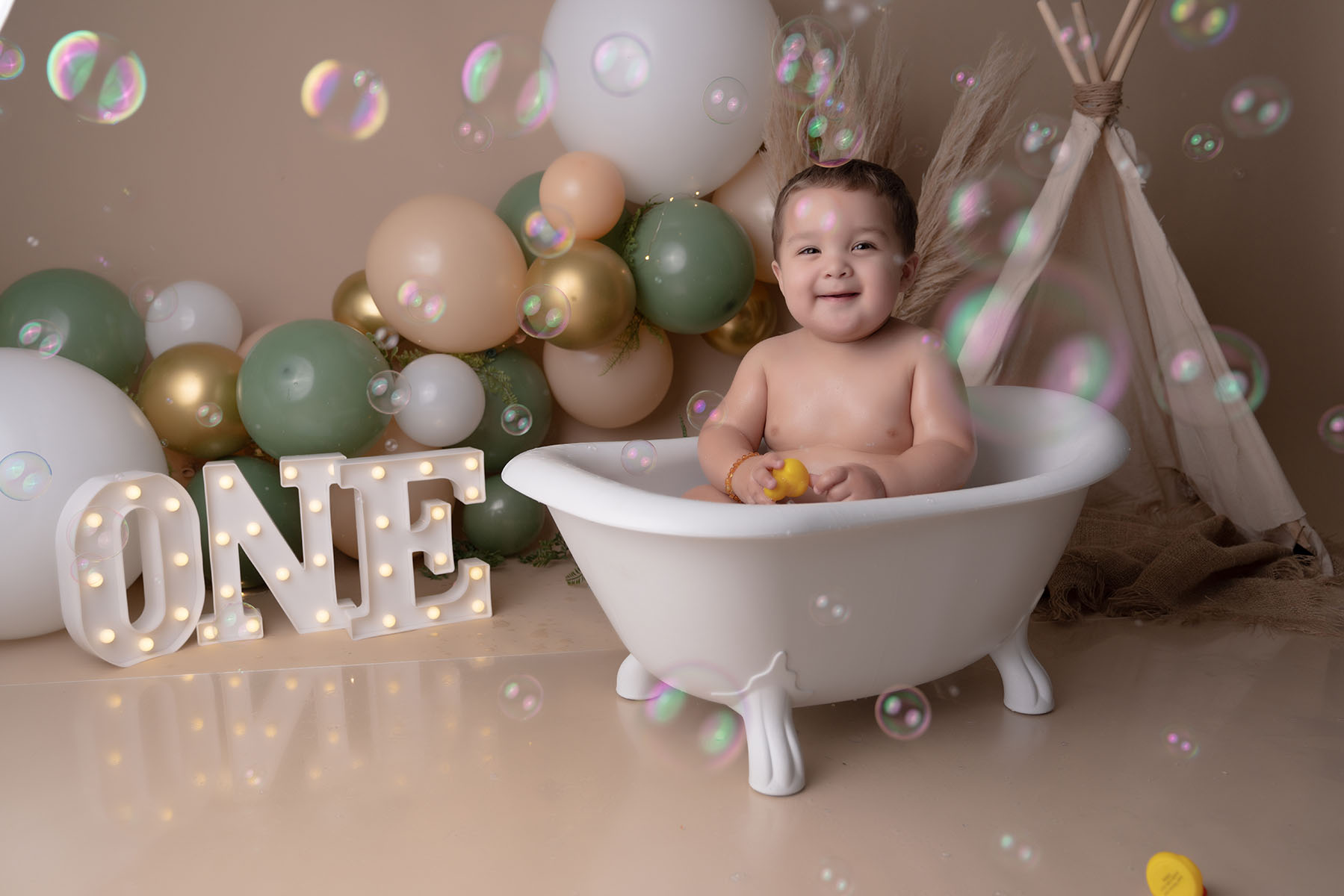 Themed newborn portrait of young boy sitting in a bathtub prop with bubbles in the air and balloons with an illuminated "One" sign in the background