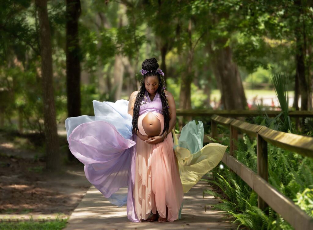 Pregnant woman in a flowing pastel dress stands on a wooded path, embracing her belly. Surrounded by greenery, she exudes serenity and joy.