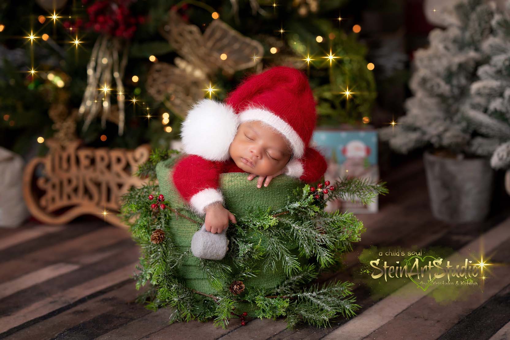 A sleeping baby wearing a Santa hat rests peacefully in a green basket adorned with festive greenery. The warm Christmas setting includes a tree, lights, and a “Merry Christmas” sign, creating a cozy holiday ambiance.