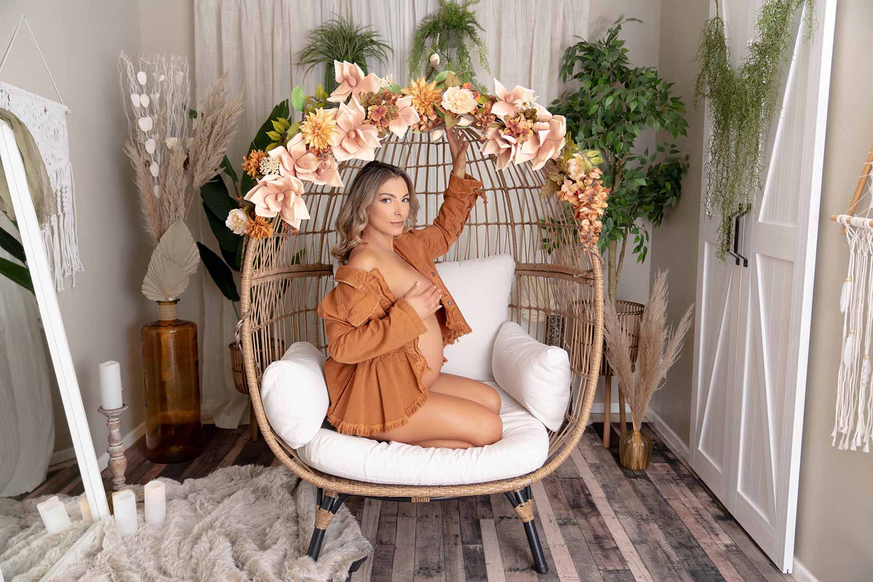 Maternity portrait of woman posed kneeling in a bohemian style chair with flowers earing a long sleeve orange dress shirt