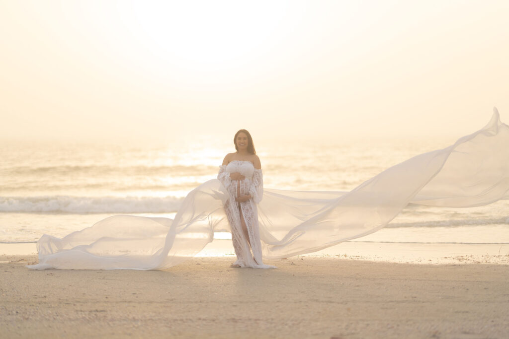 Sunset maternity portrait of woman in a flowing white gown on the beach