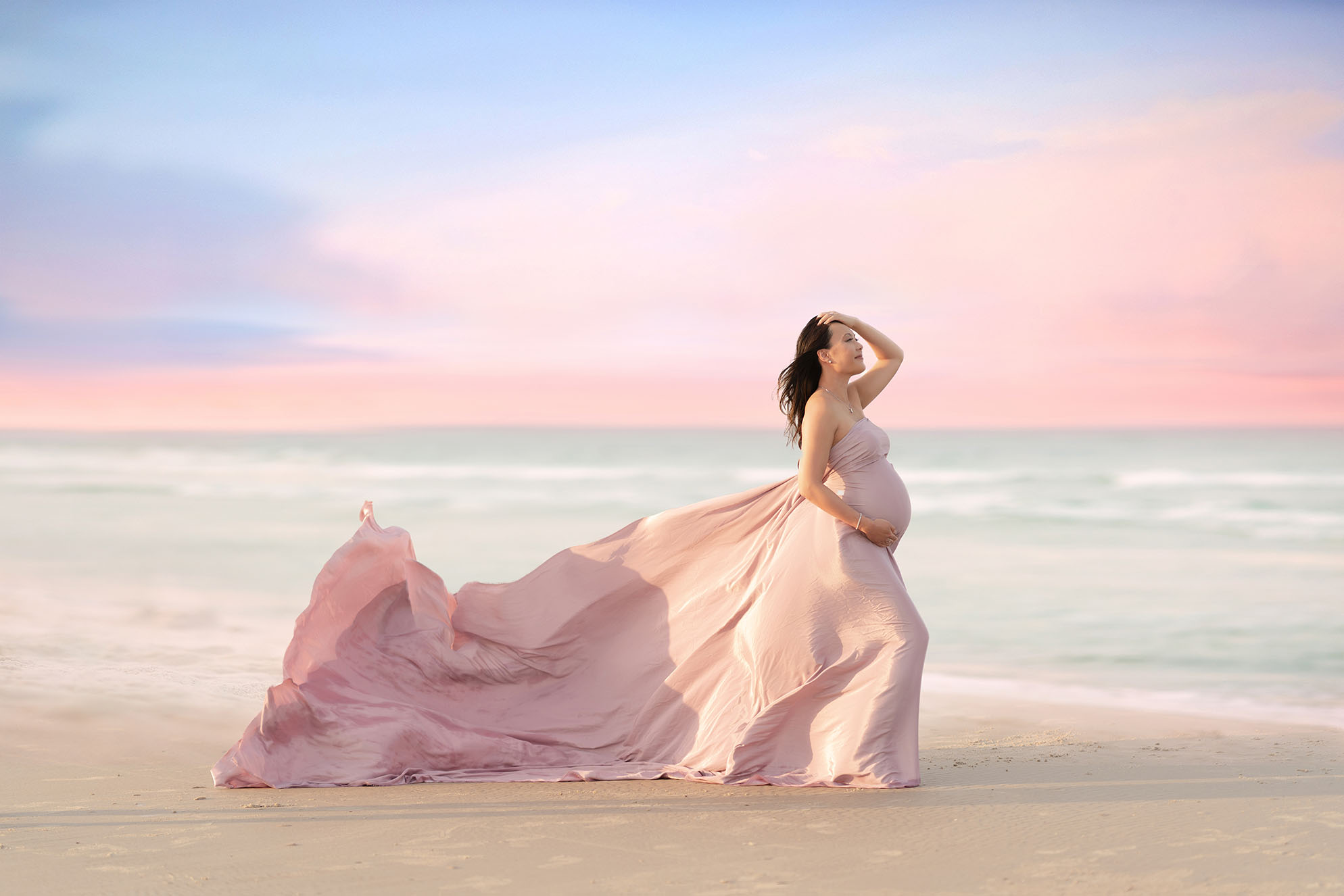 Maternity portrait of woman on a beach with a flowing gown and the sunset in the background