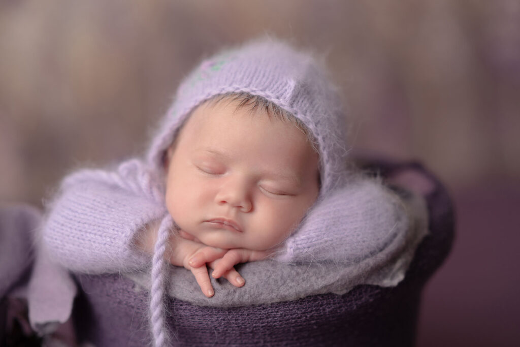 Serene newborn portrait of baby girl wearing a purple knit head covering sleeping in a pot
