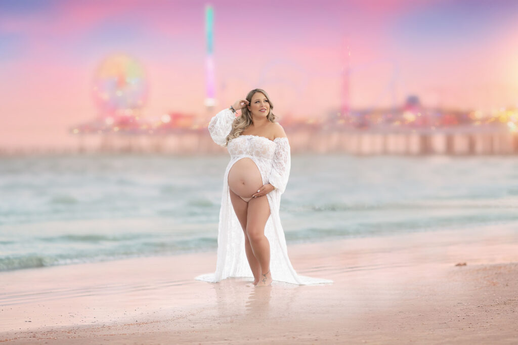 Maternity beach session of woman posed in a white gown in front of a pink sunset