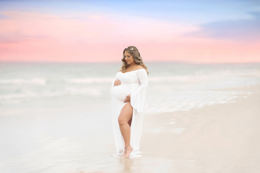 Maternity portrait of woman in a flowing white gown and flower crown at the beach with a sunset in the background