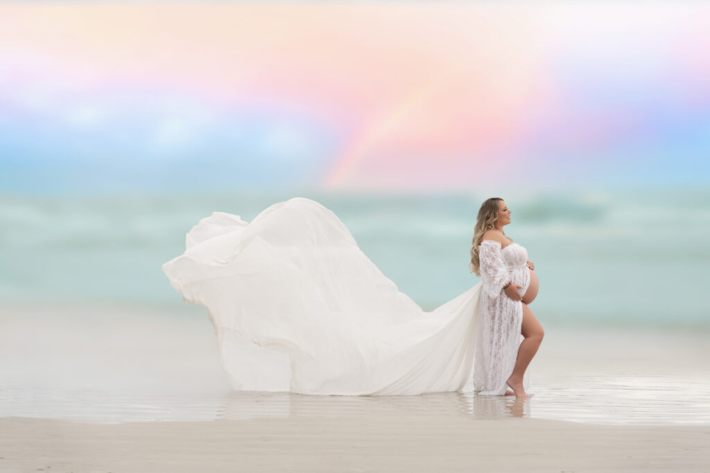 Luxury maternity portrait of blonde woman wearing a white flowing gown in front of the ocean and sunset