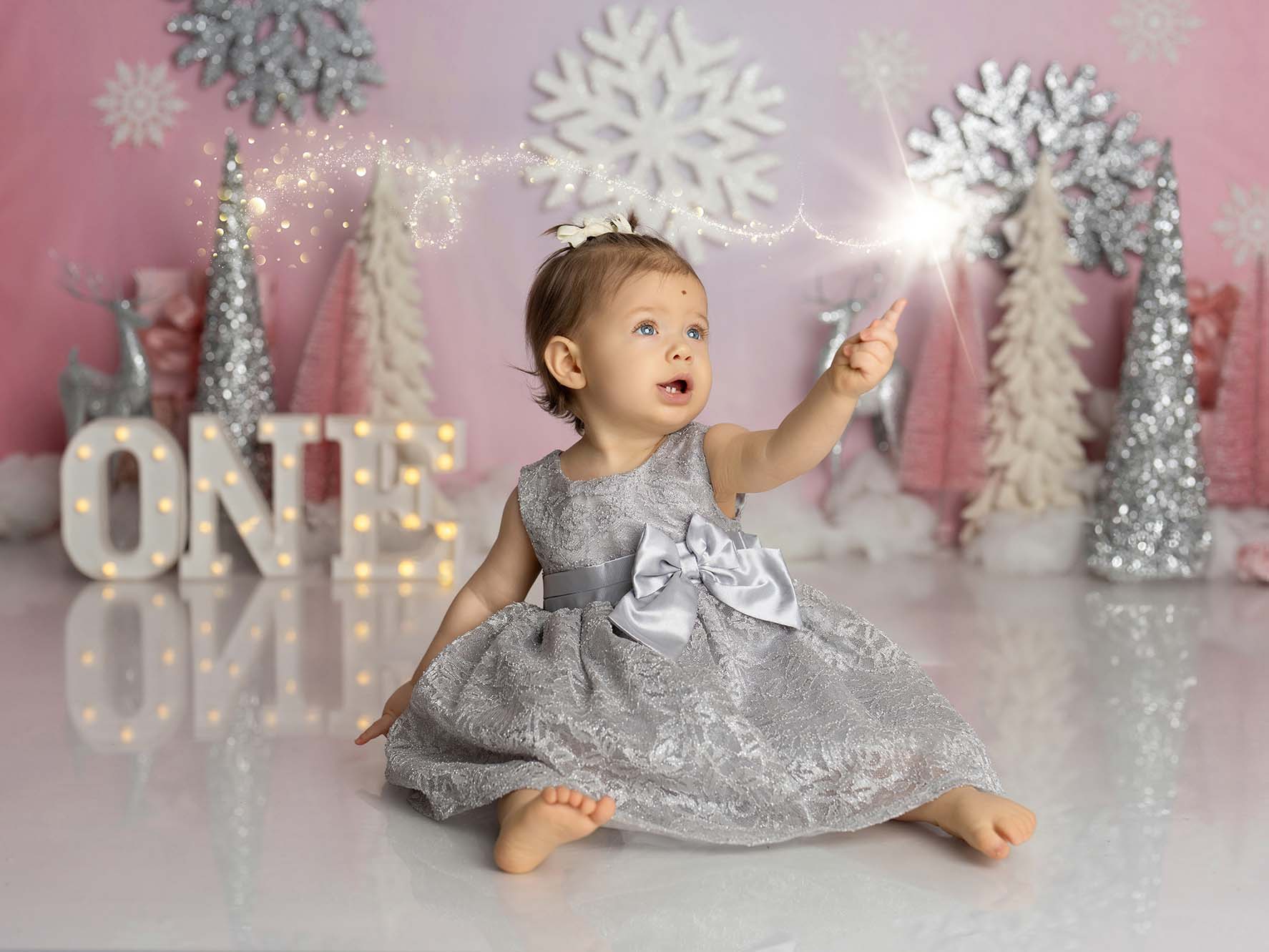 Children's portrait of young girl in a silver dress sitting in a set with snowflakes, frosted tree props and an illuminated sign spelling "One"