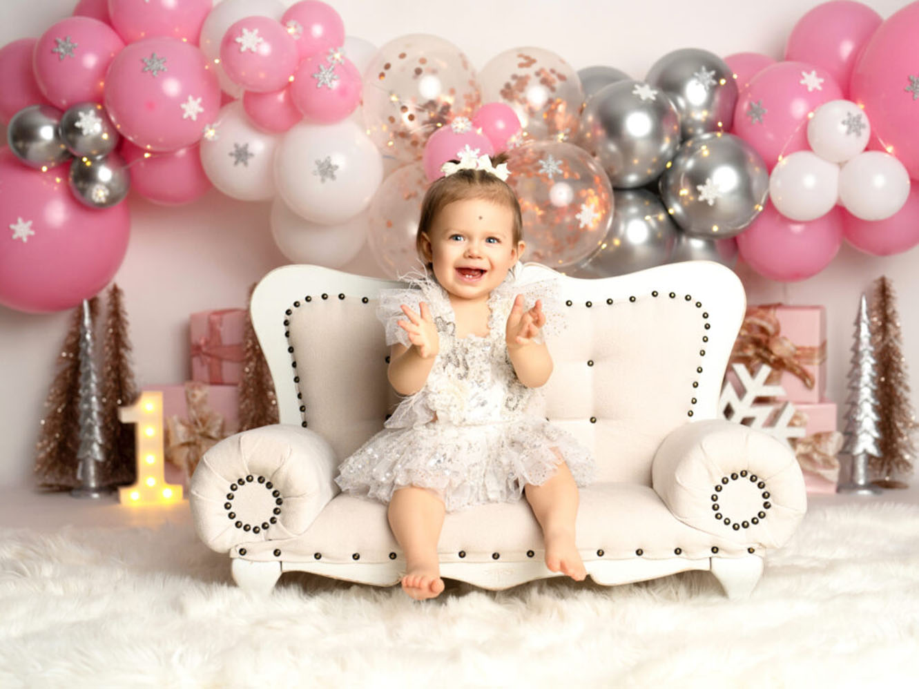 A joyful baby in a frilly dress sits on a plush white couch, surrounded by pink and silver balloons. A glowing number one signifies a celebration.