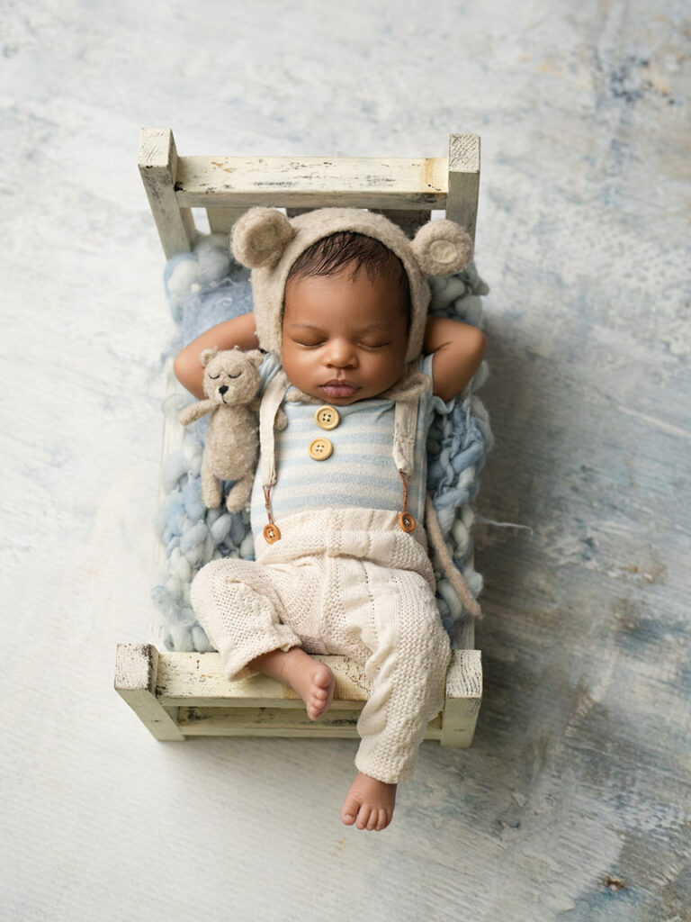A baby sleeps peacefully on a small rustic wooden bed, wearing a knitted bear hat and cream outfit, holding a stuffed bear toy. The scene is calm and cozy.