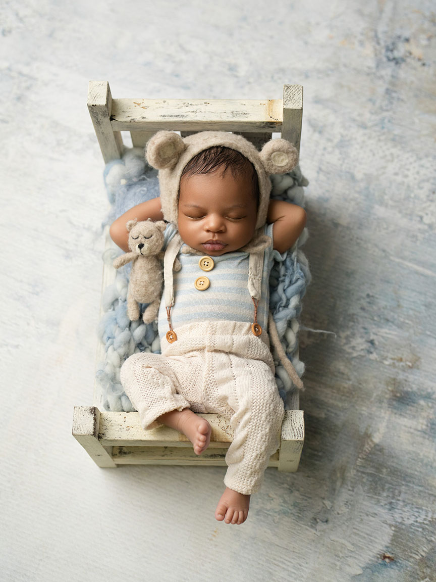 A baby sleeps peacefully on a small rustic wooden bed, wearing a knitted bear hat and cream outfit, holding a stuffed bear toy. The scene is calm and cozy.